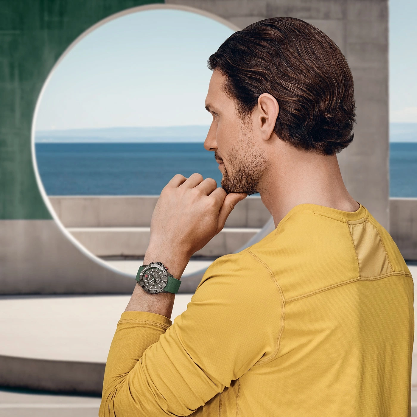 Man with watch contemplating the sea in a yellow shirt, outdoors.
