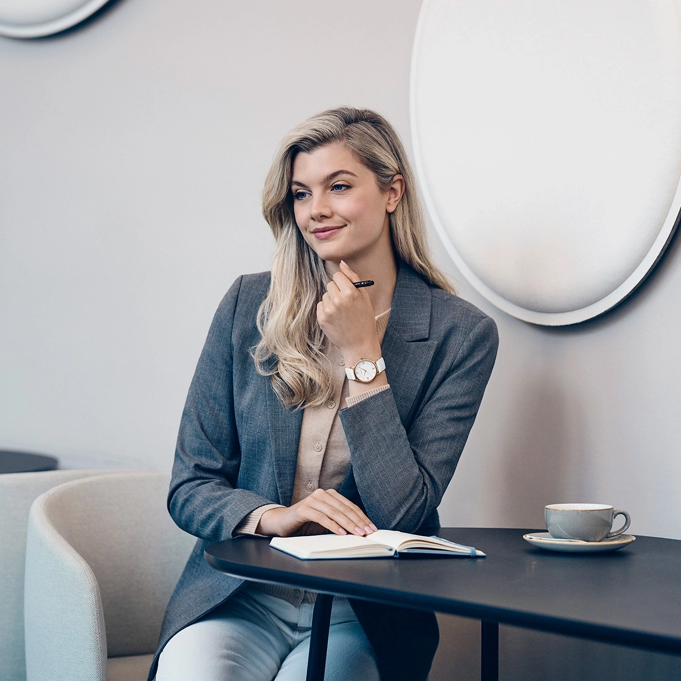 Eine Frau mit blonden Haaren sitzt an einem Tisch mit einem Notizbuch und einer Tasse Kaffee.