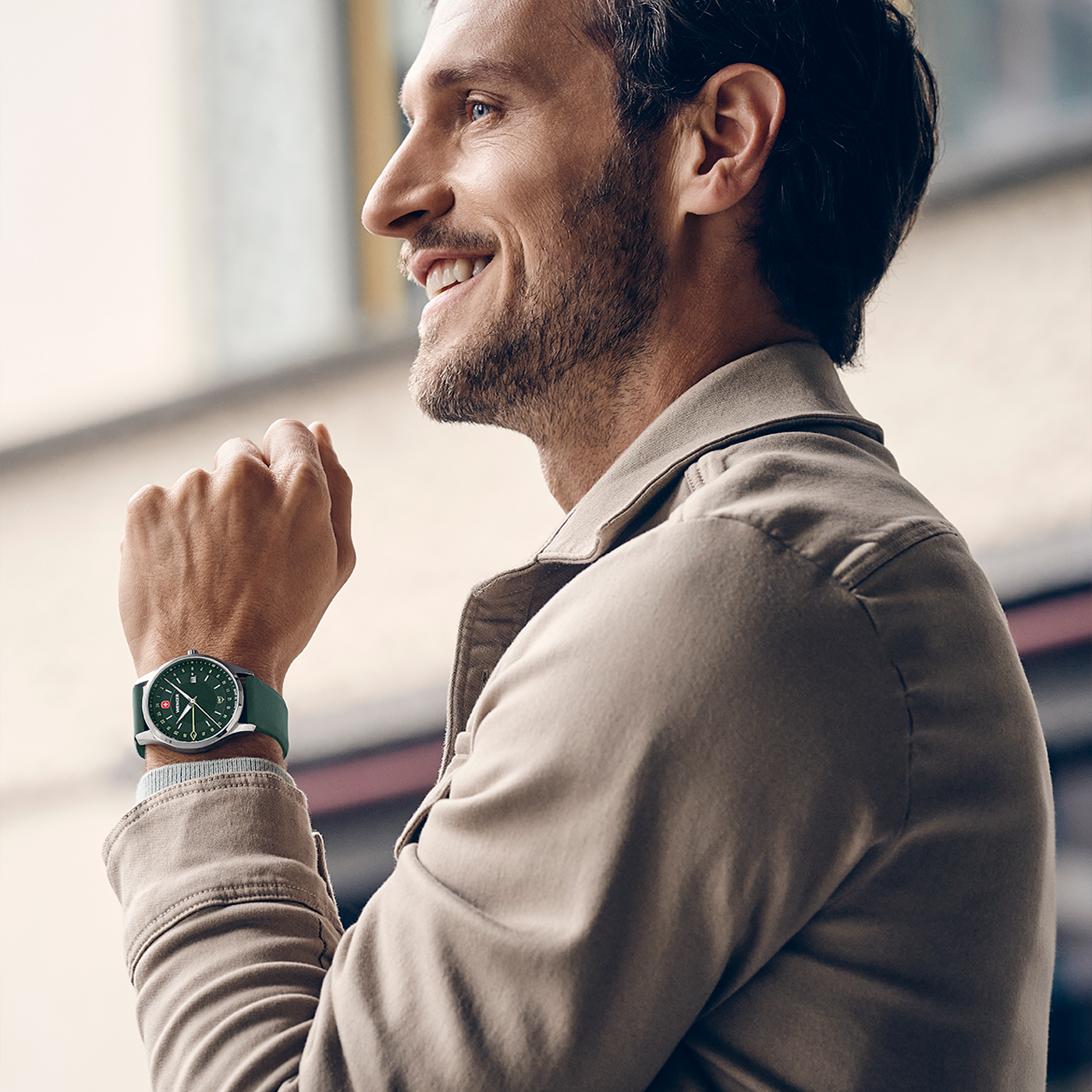 Ein Mann mit einer grünen Uhr und einem beigen Mantel lächelt.