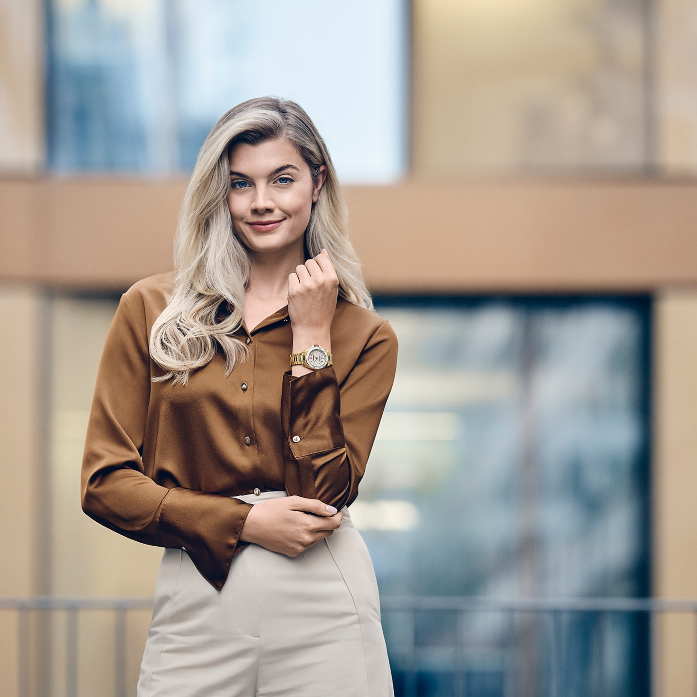 Frau mit goldenen Armbanduhr, brauner Bluse und heller Hose vor Gebäude.