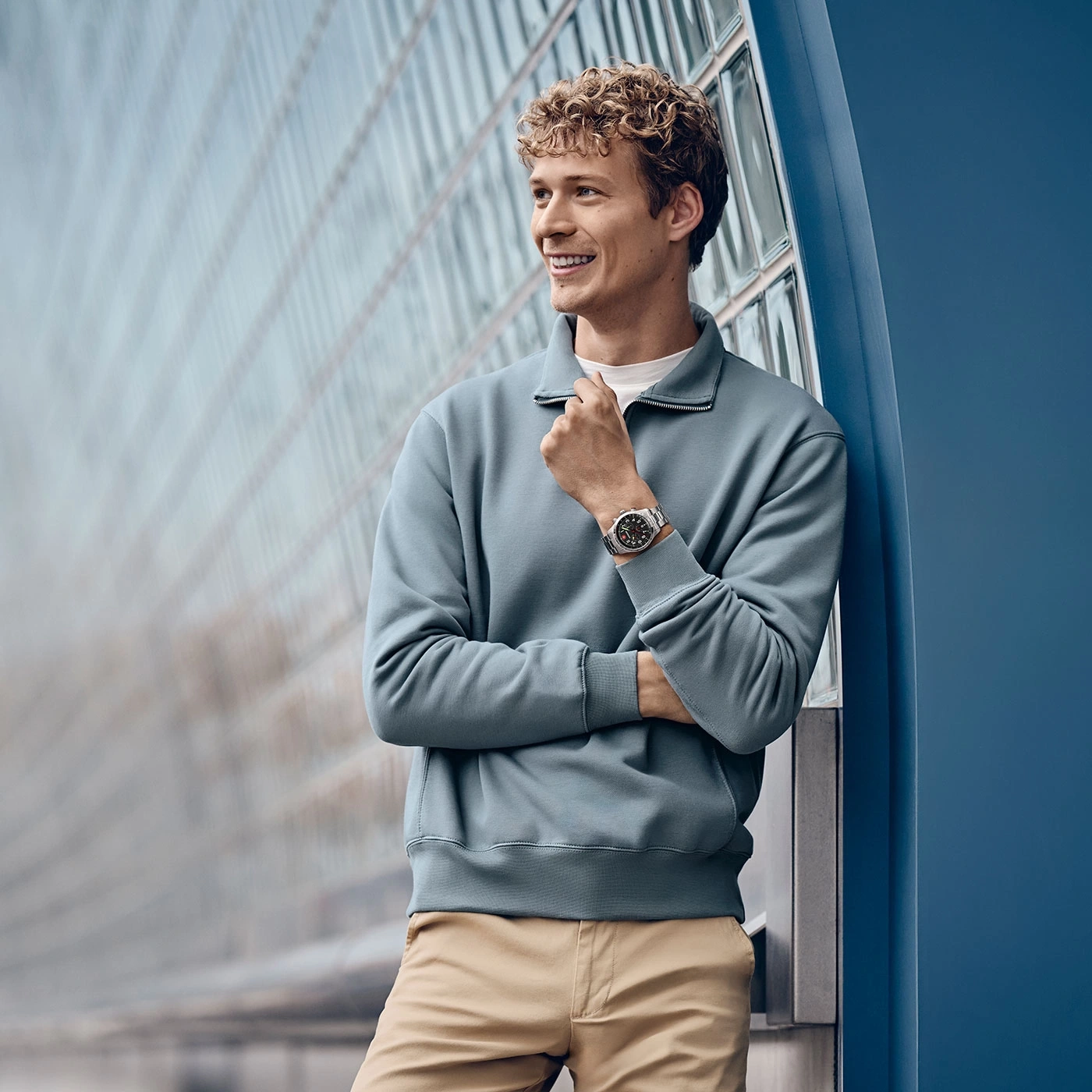 Smiling man with watch leaning against blue wall, looking away, casual outfit.