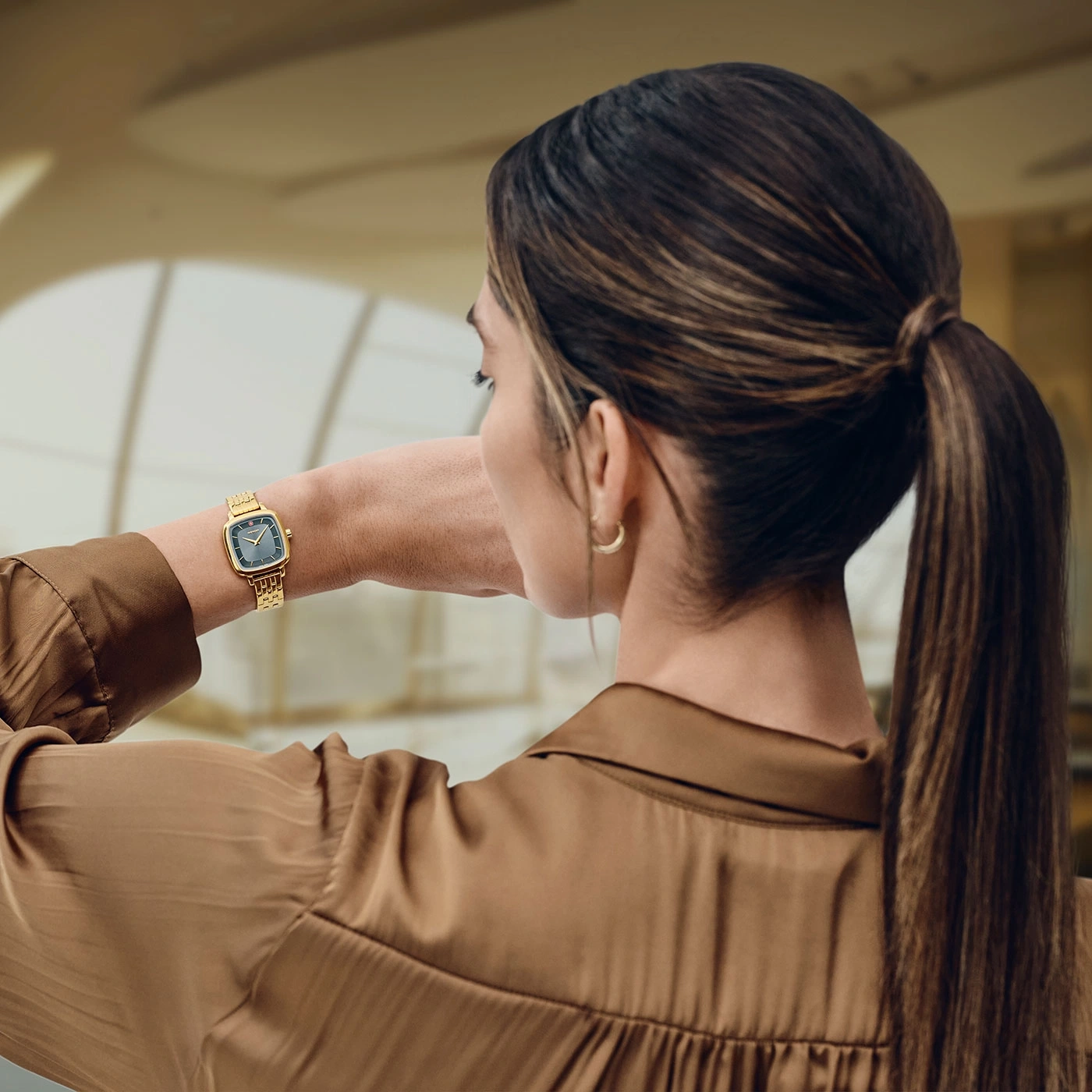 Frau mit goldener Uhr, brauner Bluse und Pferdeschwanz, Blick zur Seite
